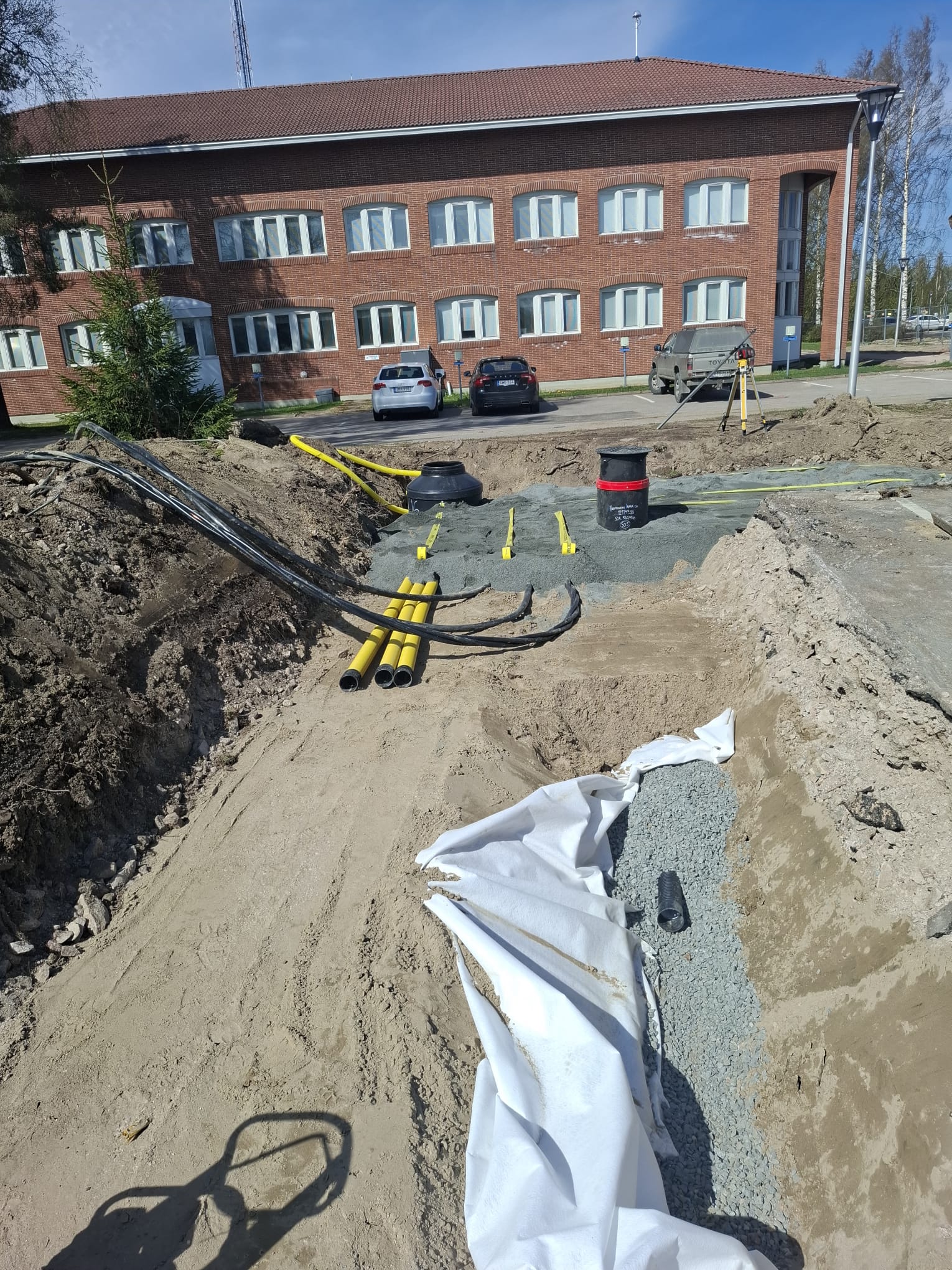 Kaivanto, jossa on keltaisia putkia, mustia kaapeleita, valkoinen suojakangas ja harmaata soraa, rakennus taustalla, aurinkoinen sää.