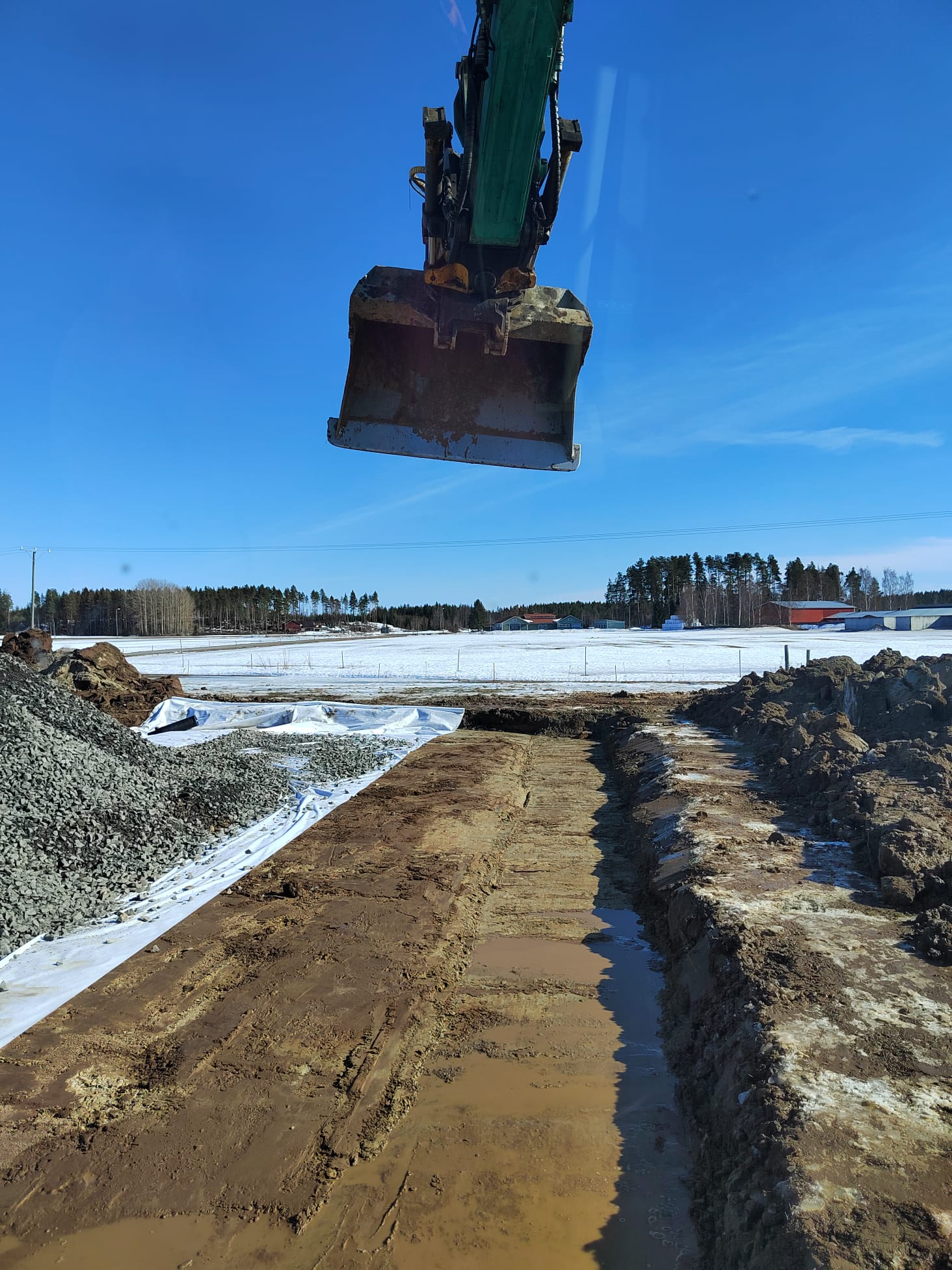 Kaivinkoneen kauha ilmassa, maaperä on mutainen ja osittain lumen peitossa, taustalla metsä ja sininen taivas.