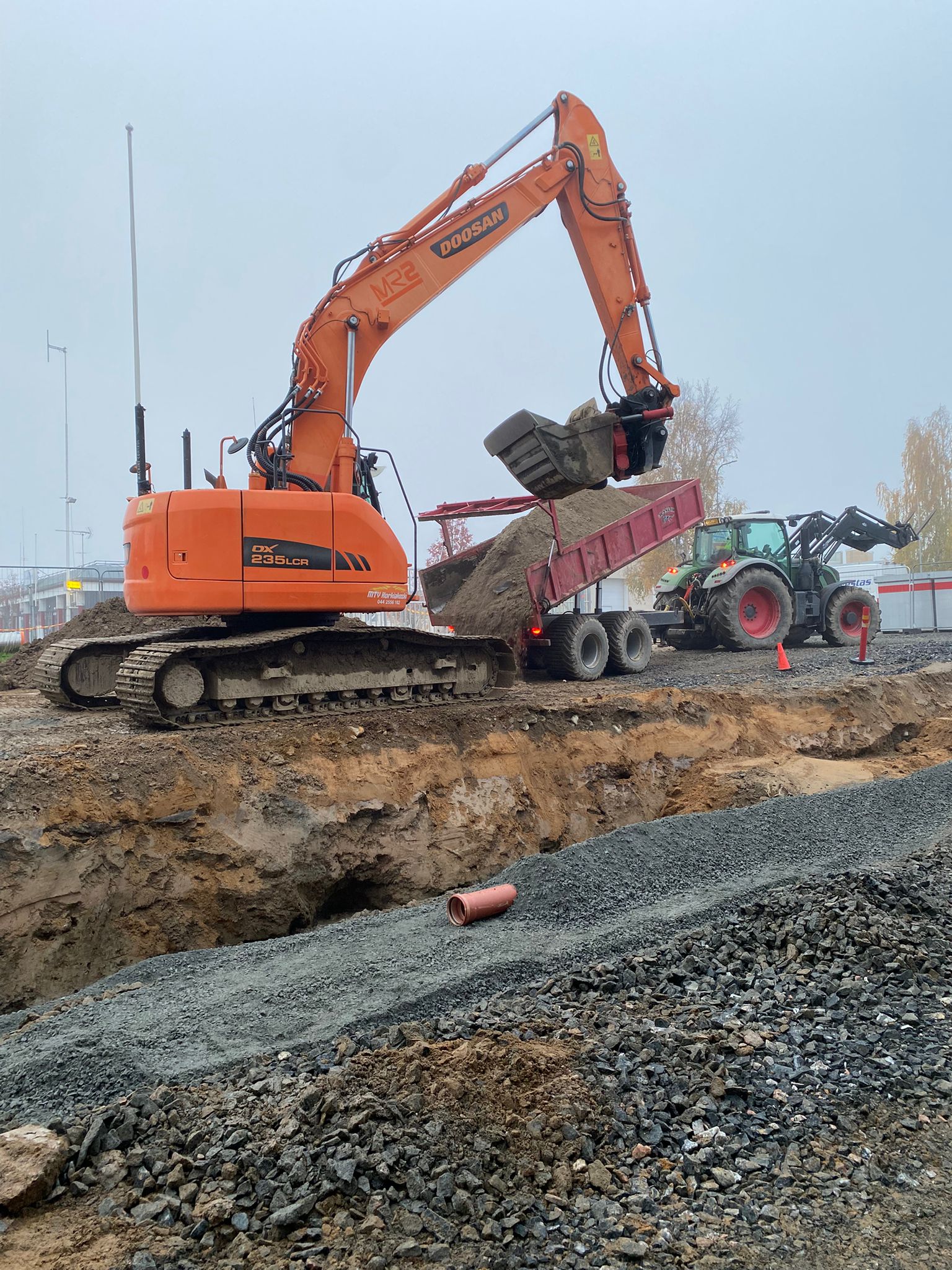 Oranssi Doosan-kaivinkone lastaa maata punaiseen perävaunuun, vihreä traktori taustalla, harmaa sora etualalla, pilvinen sää.