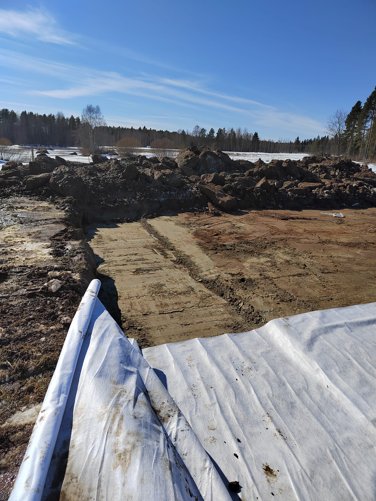 Maarakennustyömaa aurinkoisena päivänä, jossa näkyy maata, lunta, puita ja valkoinen suojakangas etualalla.