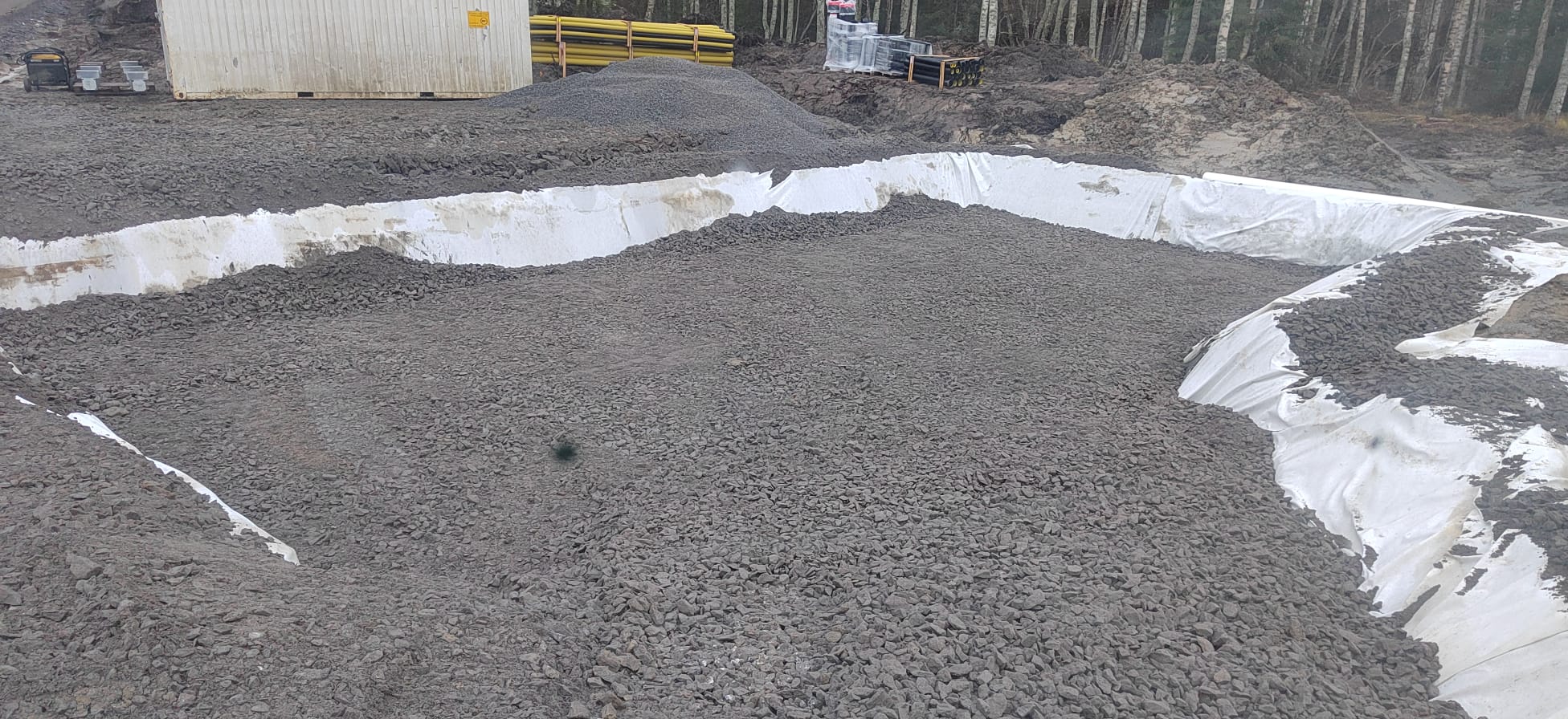 Rakennustyömaalla on harmaata mursketta valkoisen suojakankaan päällä, taustalla kontti, putkia ja puita. Valo on himmeää.