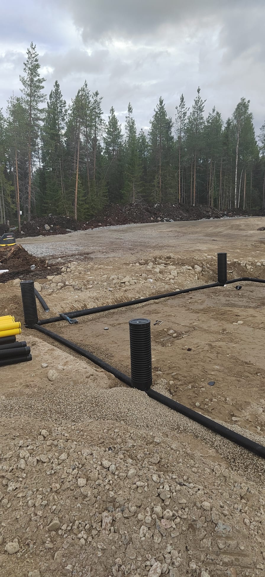 Maaperässä on mustia ja keltaisia putkia, jotka muodostavat neliön, taustalla on puita ja harmaa taivas.