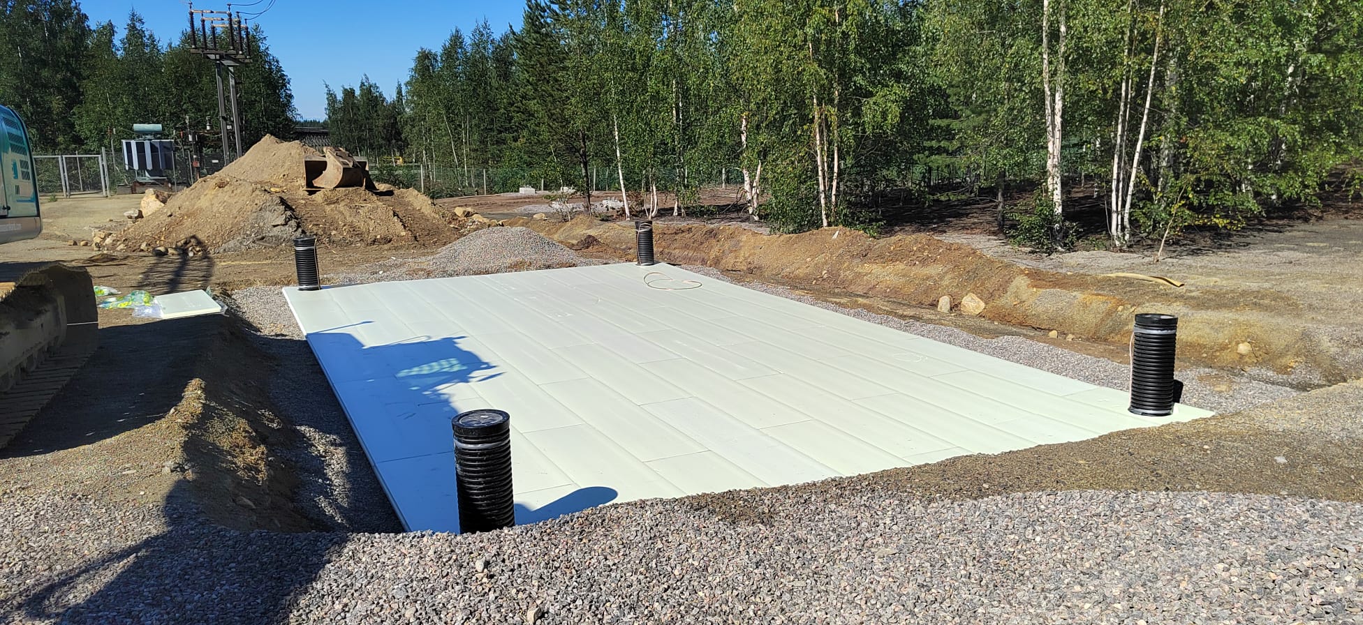 Rakennustyömaa, jossa on valkoinen alusta, mustat putket ja harmaa kivimurske, aurinkoisessa säässä.