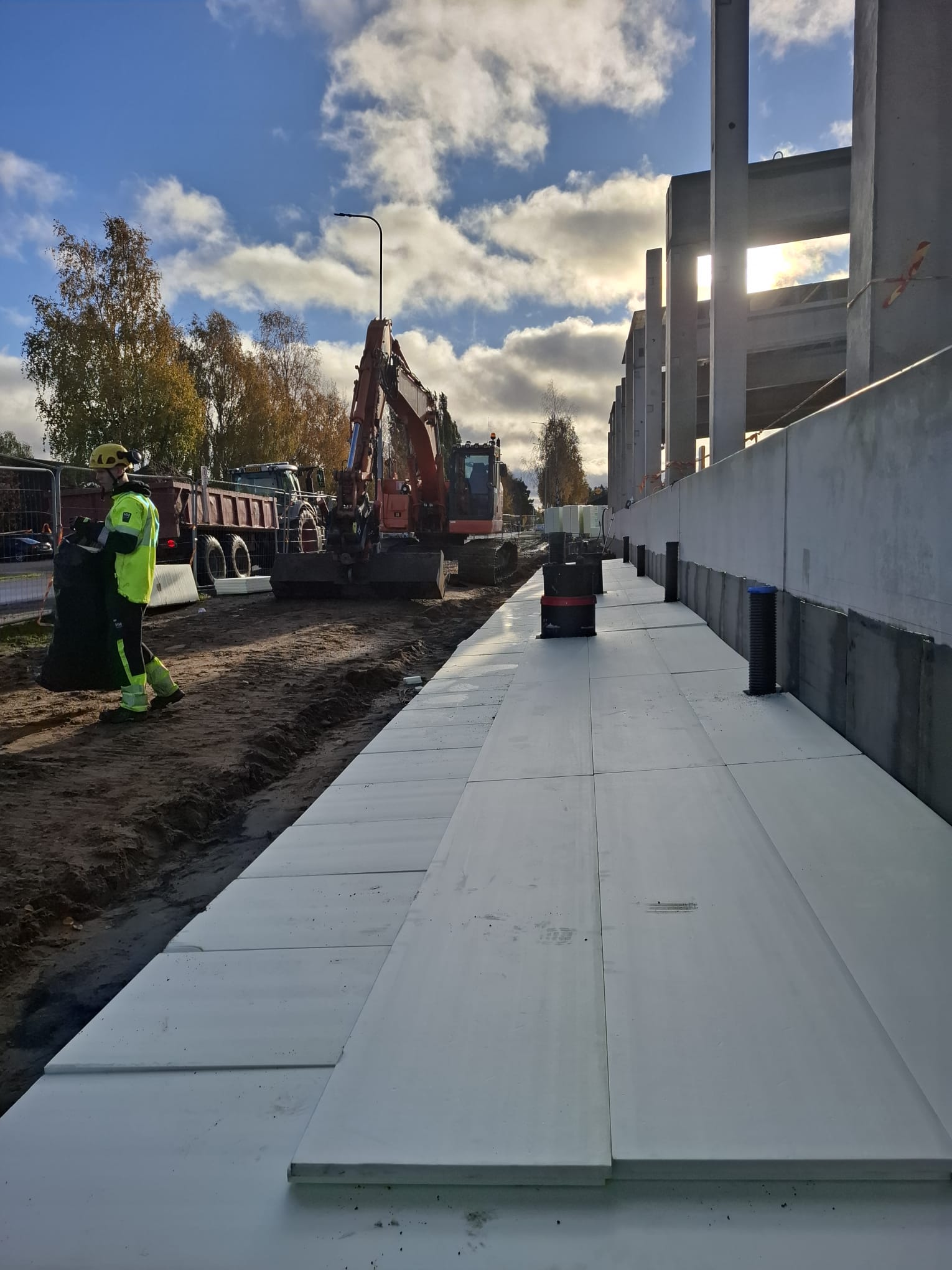 Rakennustyömaalla näkyy työntekijä suojavarusteissa, kaivinkone, kuorma-auto ja valkoisia betonilaattoja auringonpaisteessa.