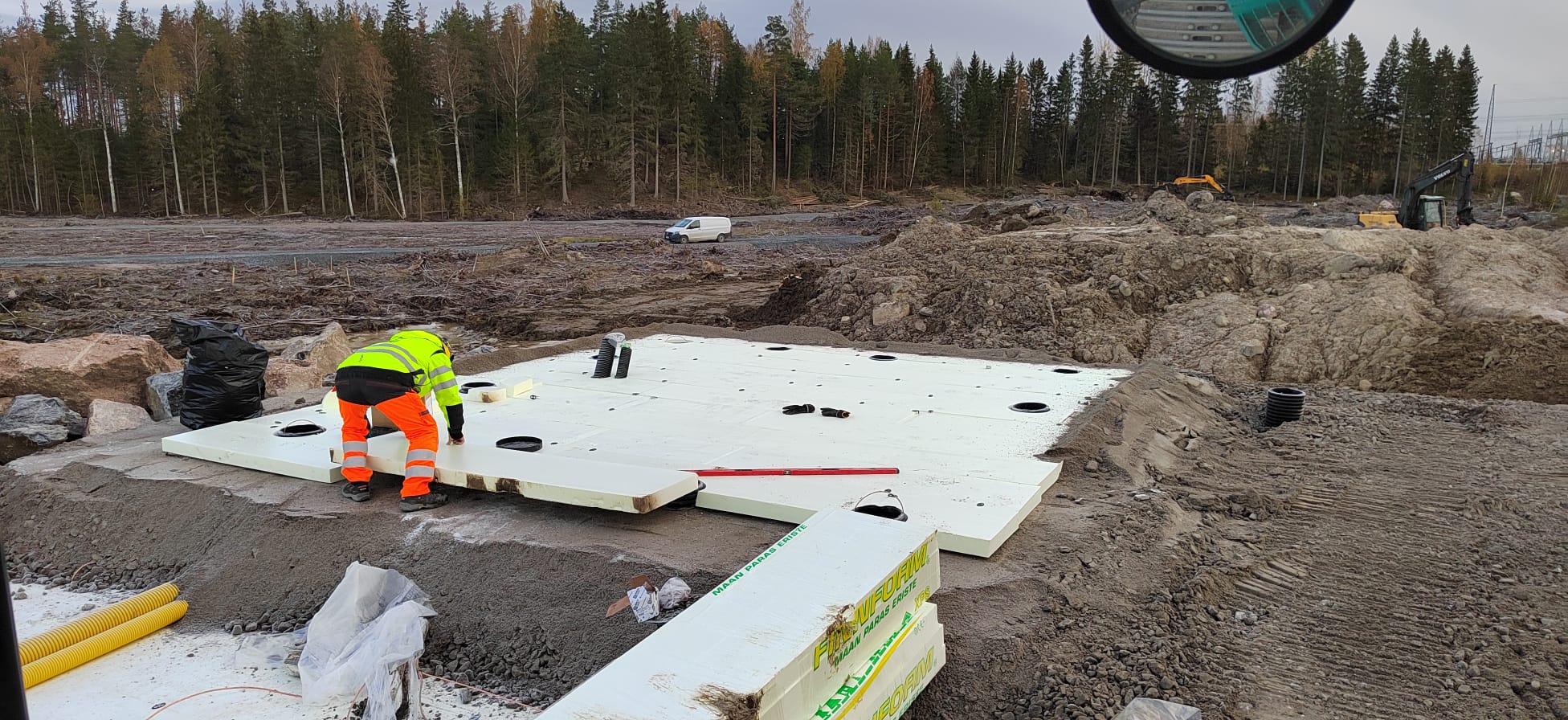 Rakennustyömaalla työskentelee henkilö, joka asentaa valkoisia eristyslevyjä maahan, taustalla näkyy metsää ja kaivinkoneita, valaistus on luonnollinen ja pilvinen.