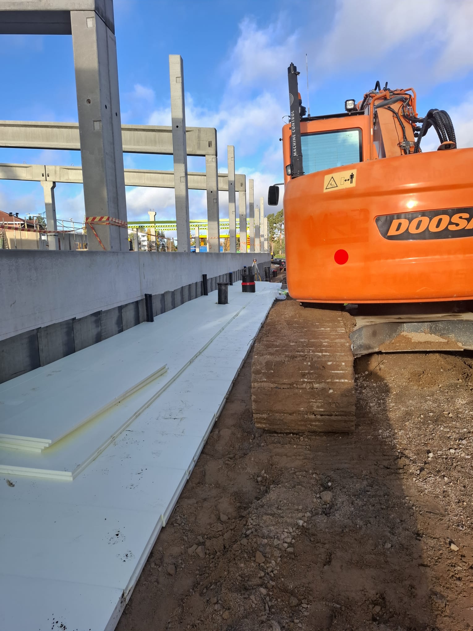 Rakennustyömaa, jossa on oranssi Doosan-kaivinkone, valkoista eristyslevyä ja betonipylväitä sinisen taivaan alla.