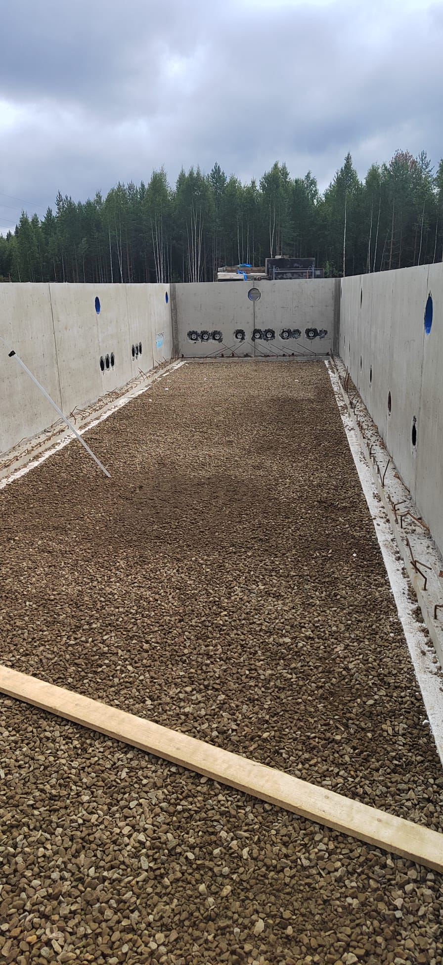 Rakennustyömaa, jossa on kivillä täytetty betonirakenne, harmaa taivas ja metsä taustalla.