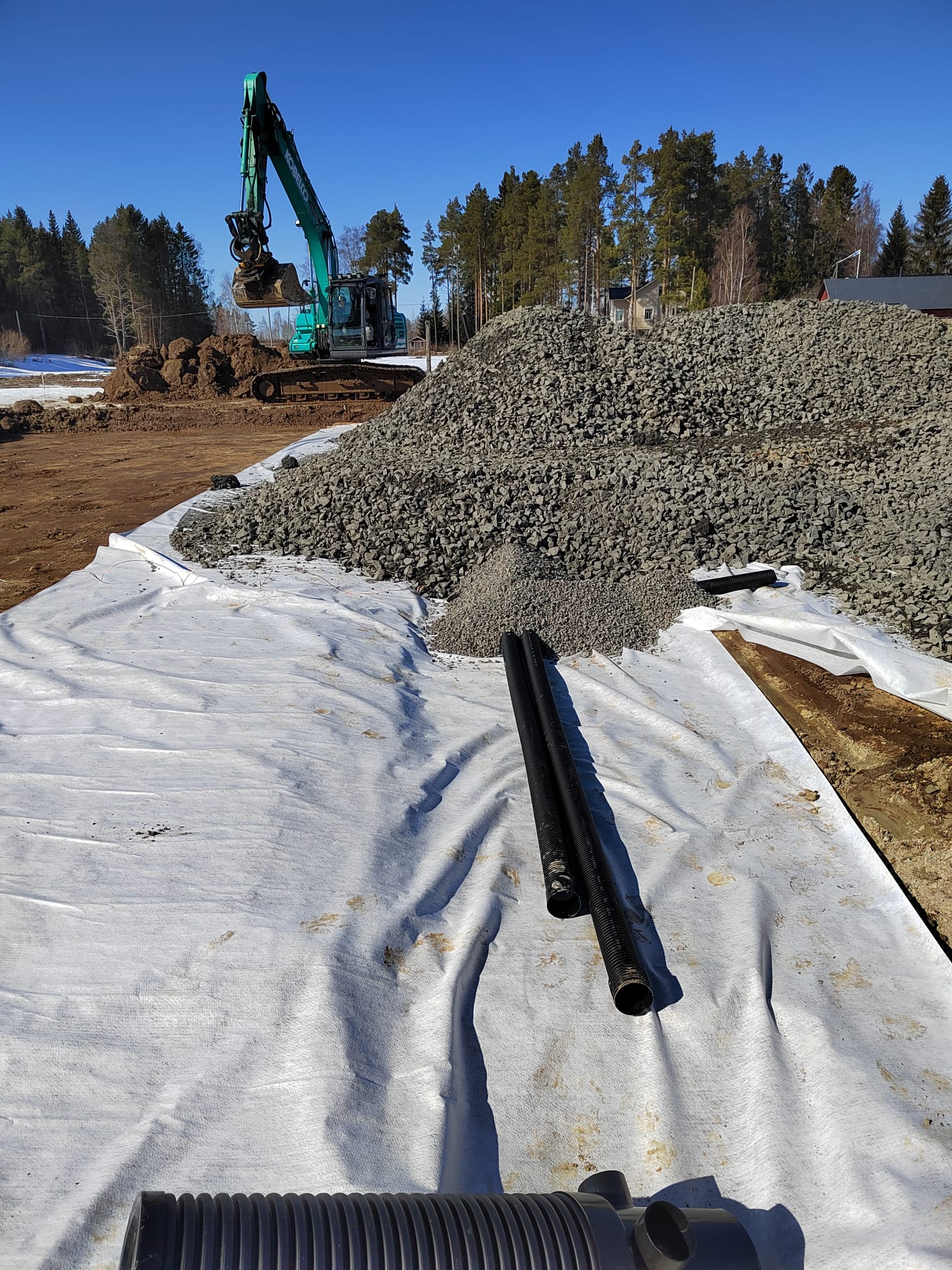 Rakennustyömaalla on suuri kivikasa valkoisella suojakankaalla, kaivinkone taustalla ja mustia putkia etualalla, aurinkoisessa säässä.