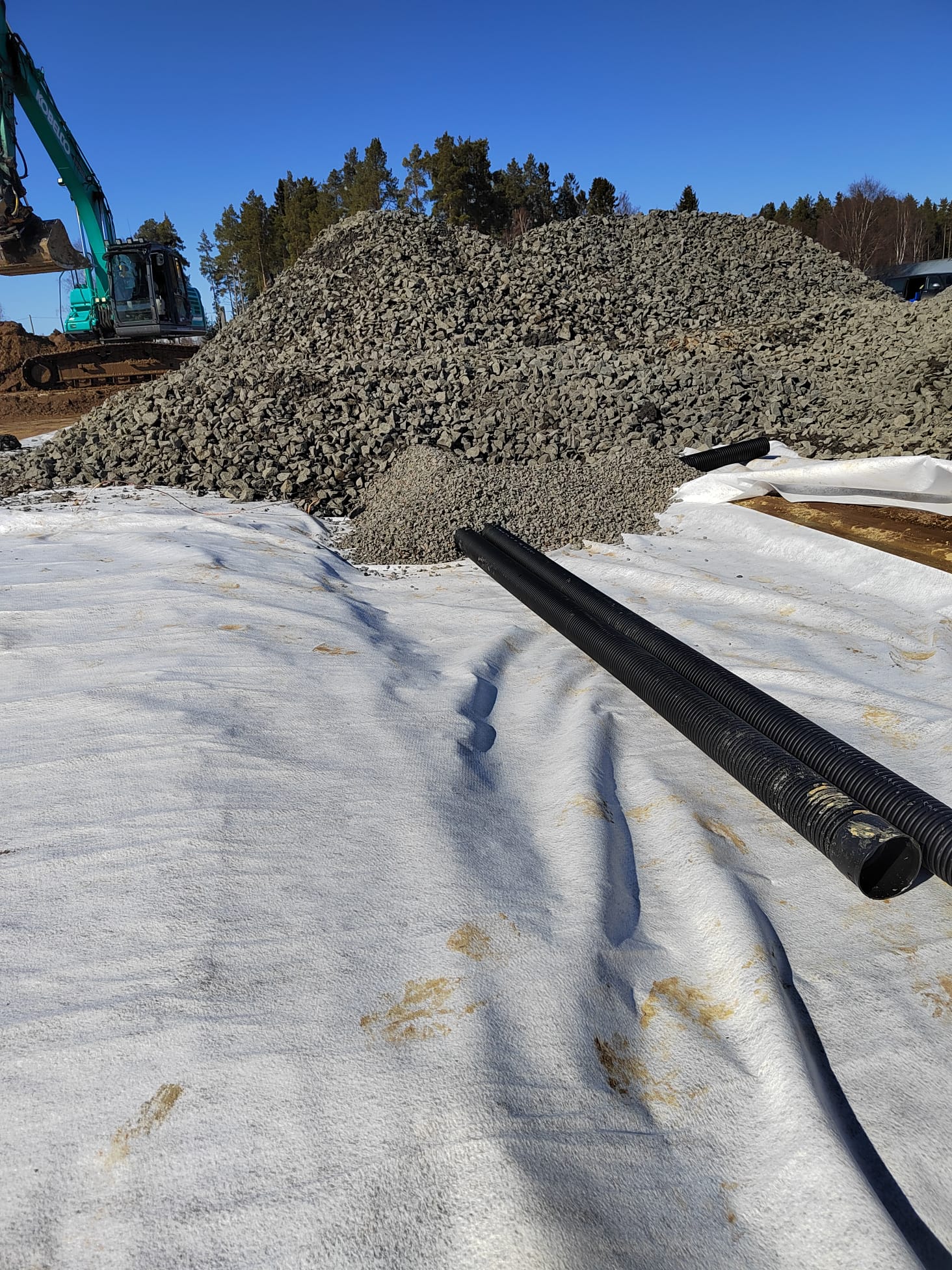 Rakennustyömaalla on valkoinen suodatinkangas, jonka päällä on kaksi mustaa putkea ja suuri kivikasa taustalla, aurinkoisessa säässä.