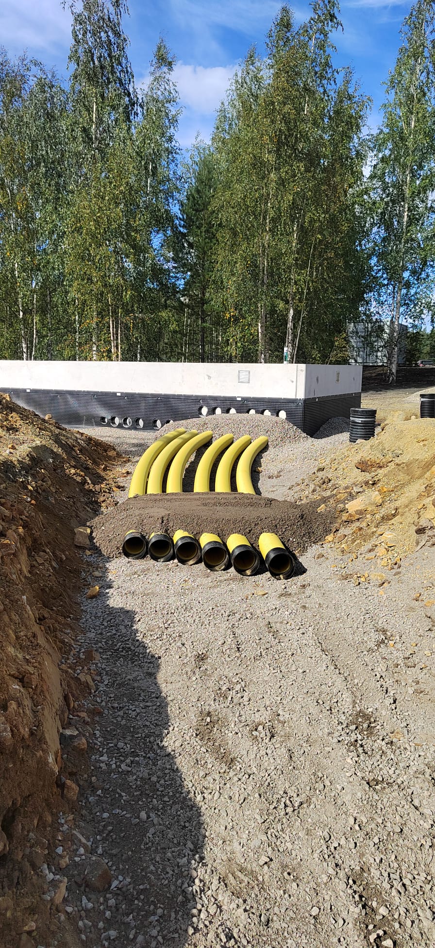 Ulkona on rakennustyömaa, jossa näkyy keltaisia putkia, kivituhkaa ja maata, sekä taustalla puita ja sininen taivas.