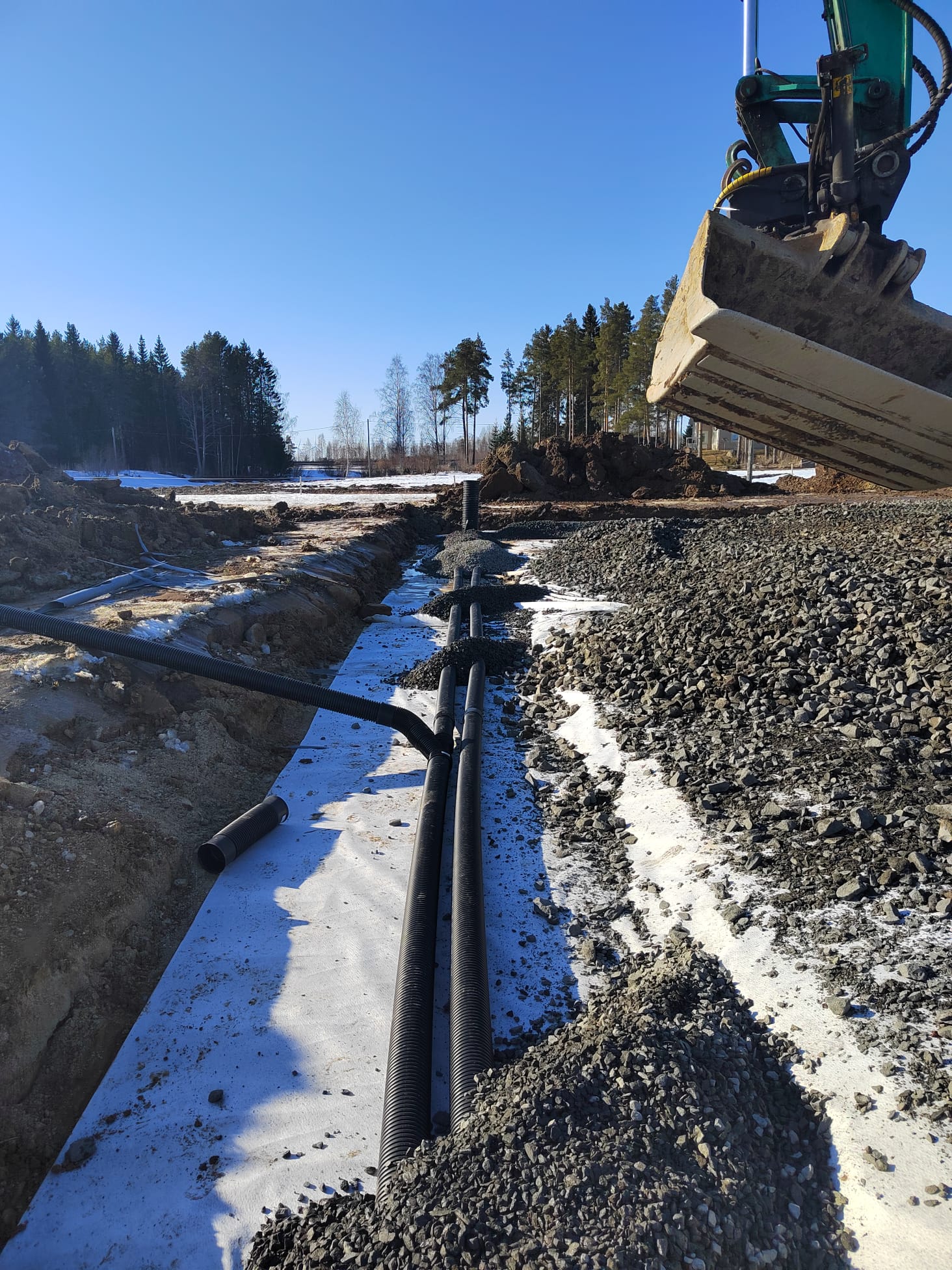 Rakennustyömaa, jossa on mustia putkia valkoisella kankaalla, kivikkoa ja kaivinkoneen kauha ylhäällä, aurinkoisena päivänä.