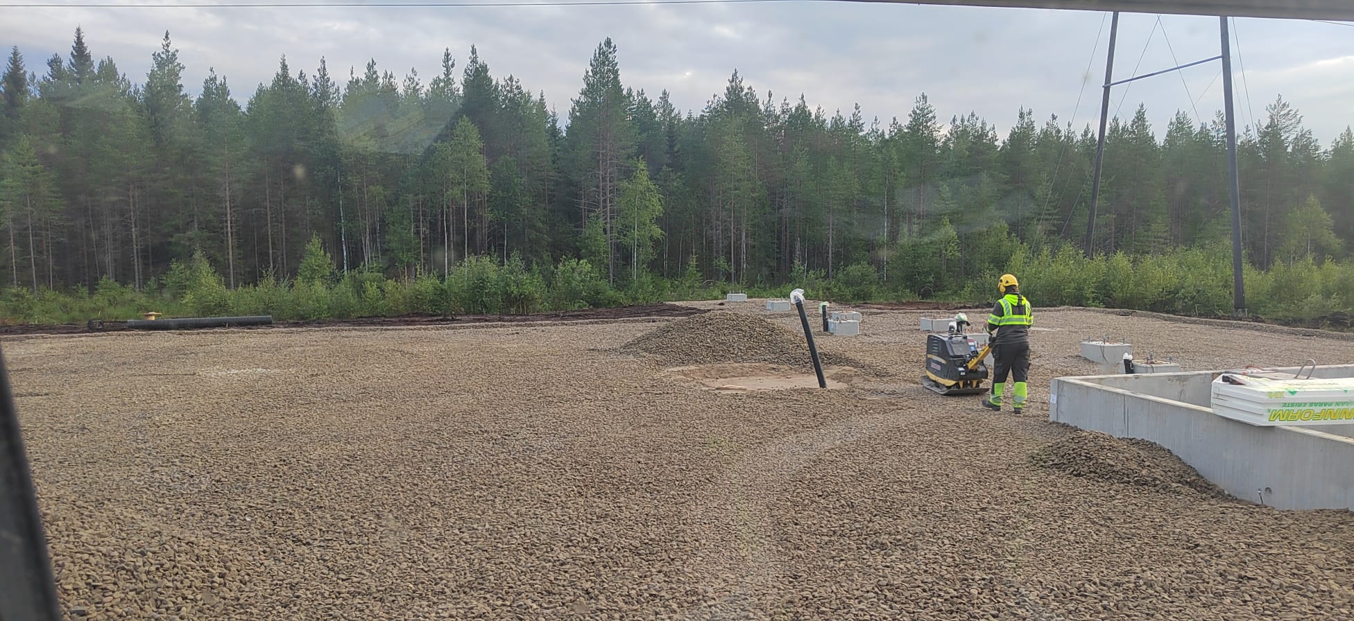 Rakennustyömaa, jossa on sorapintainen alue, betonirakenteita, työntekijä tiivistämässä maata ja taustalla metsä. Valoisa sää.