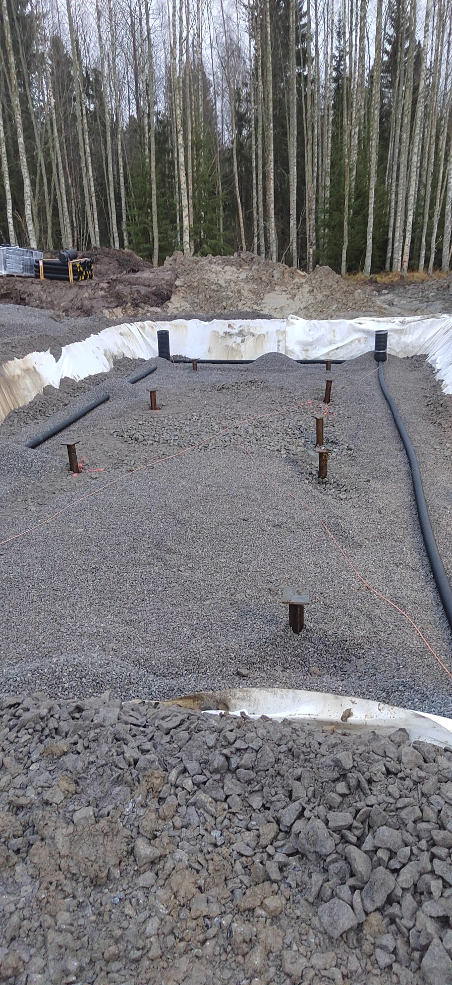 Rakennustyömaa, jossa on harmaata soraa, mustia putkia ja valkoista suojakangasta, taustalla puita ja harmaa taivas.