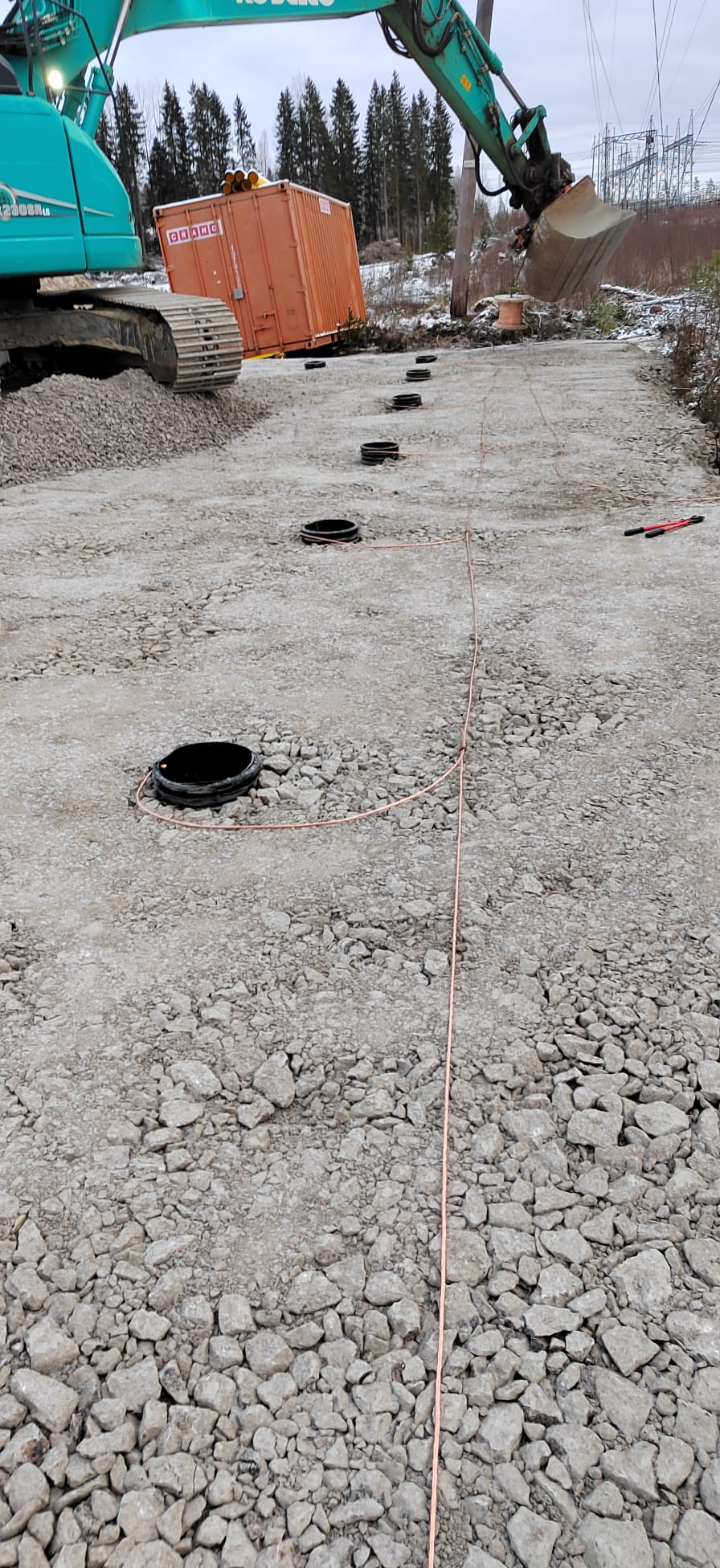 Ulkona on rakennustyömaa, jossa on kaivinkone, oranssi kontti ja useita kaivoja, jotka on yhdistetty kuparilangalla. Valoisa, pilvinen sää.