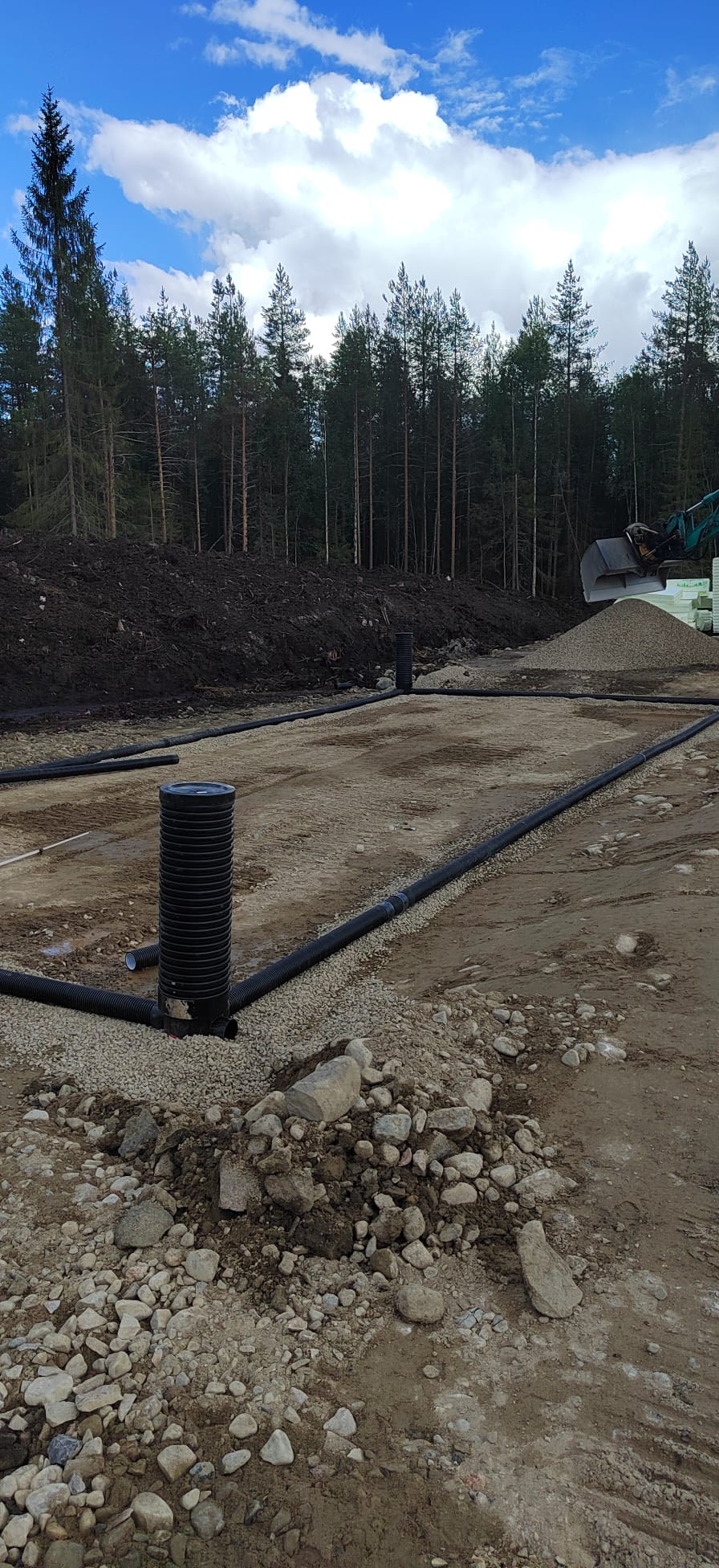 Ulkona on rakennustyömaa, jossa näkyy mustia putkia, kivikasoja, maata, puita ja sininen taivas pilvillä.