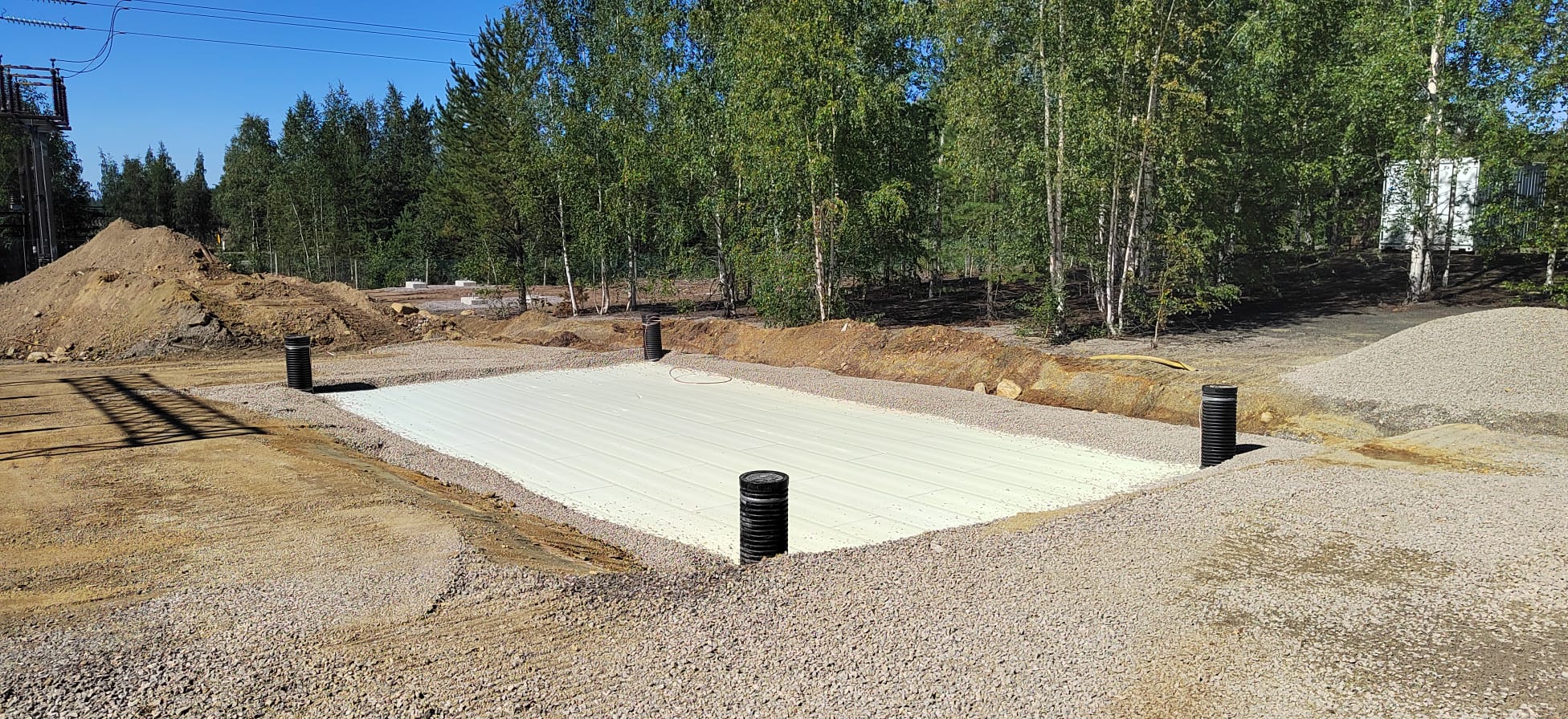 Rakennustyömaa, jossa on valkoinen pohja, mustat putket, kivimurska ja taustalla puita aurinkoisessa säässä.