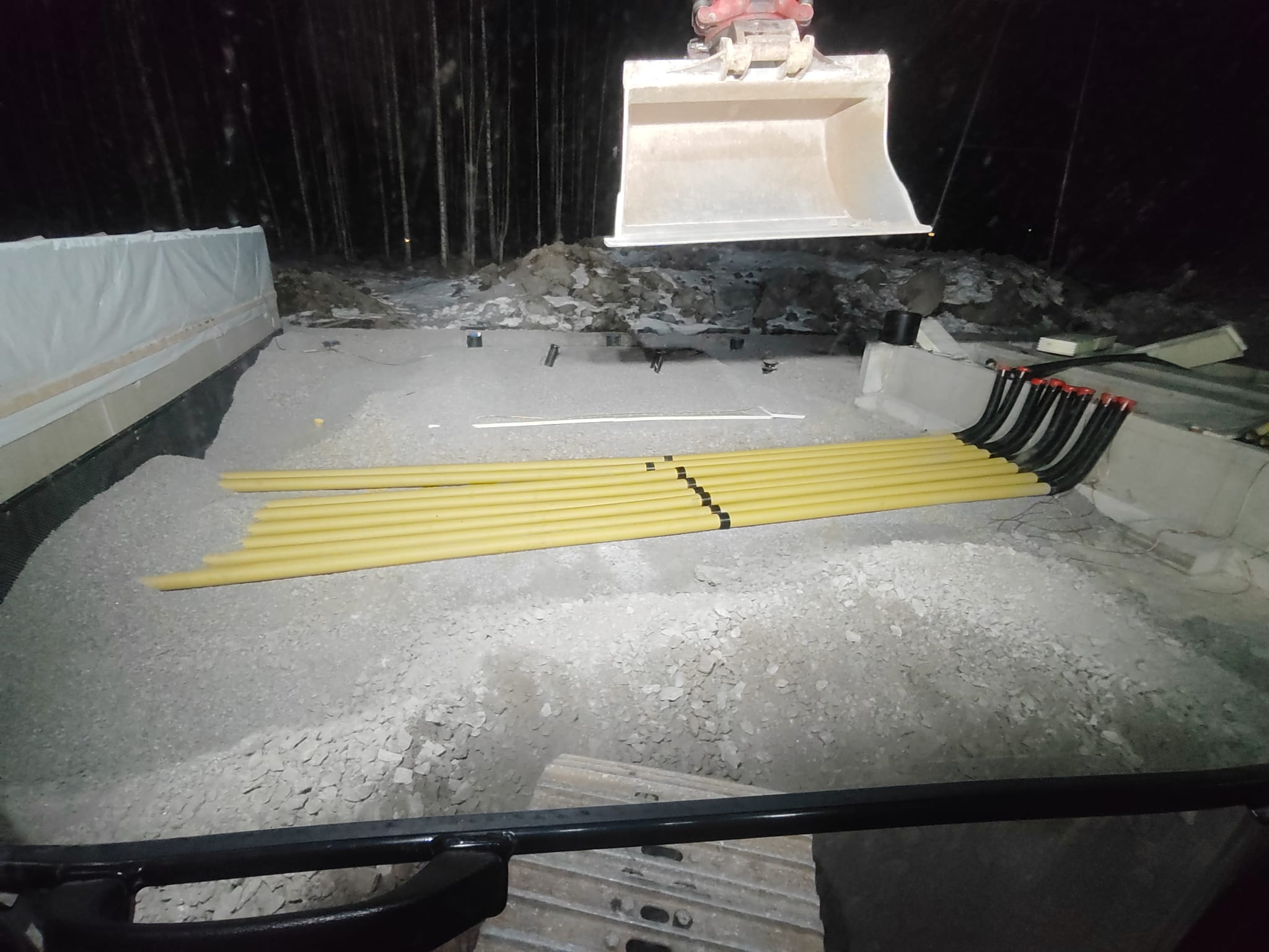 Rakennustyömaa yöllä, jossa näkyy keltaisia putkia, kaivinkoneen kauha ja sorapintaa, valaistus on heikkoa.