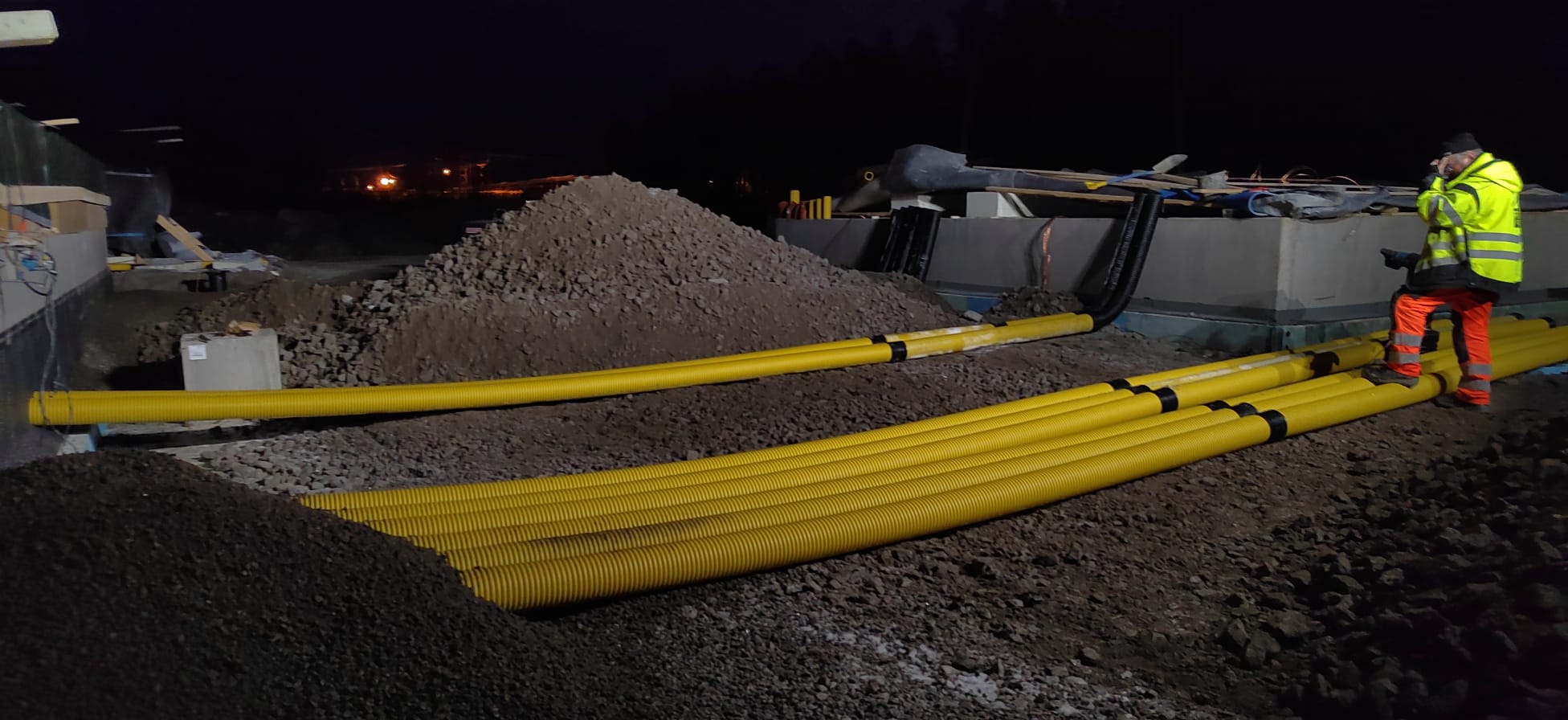 Yöllinen rakennustyömaa, jossa näkyy keltainen putkisto, maakasa ja työntekijä huomiovaatteissa. Valot ovat himmeät.