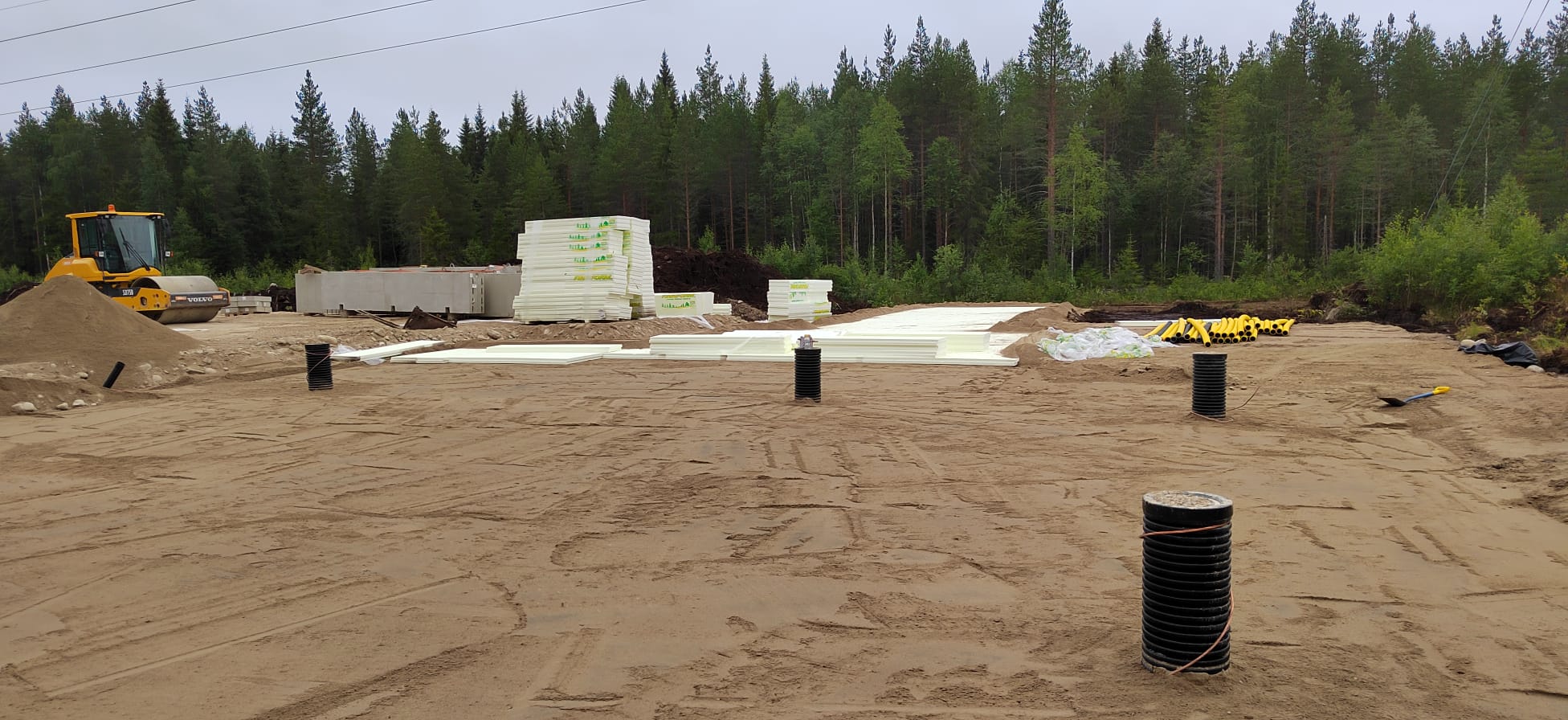 Rakennustyömaa, jossa on keltainen jyrä, valkoisia eristelevyjä, mustia putkia ja hiekkaa, metsän reunalla pilvisenä päivänä.