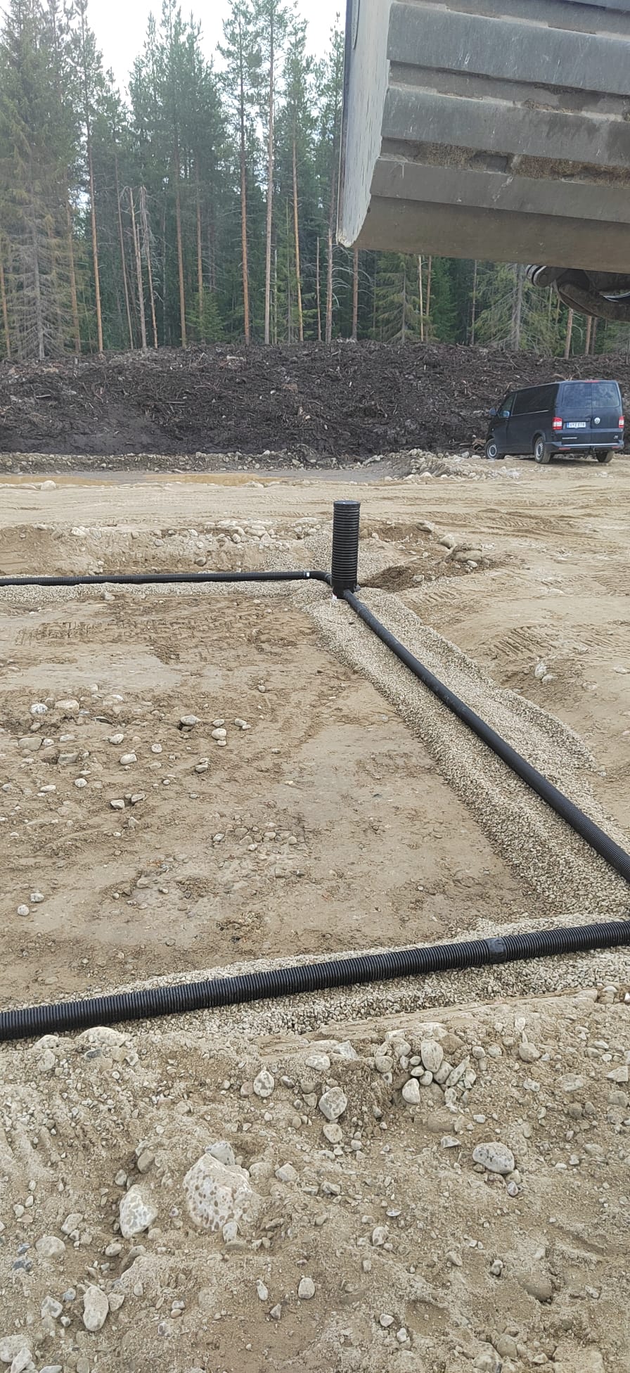Maaperään asennettu musta salaojaputki, jossa on harmaa sora, taustalla metsä ja tumma pakettiauto.