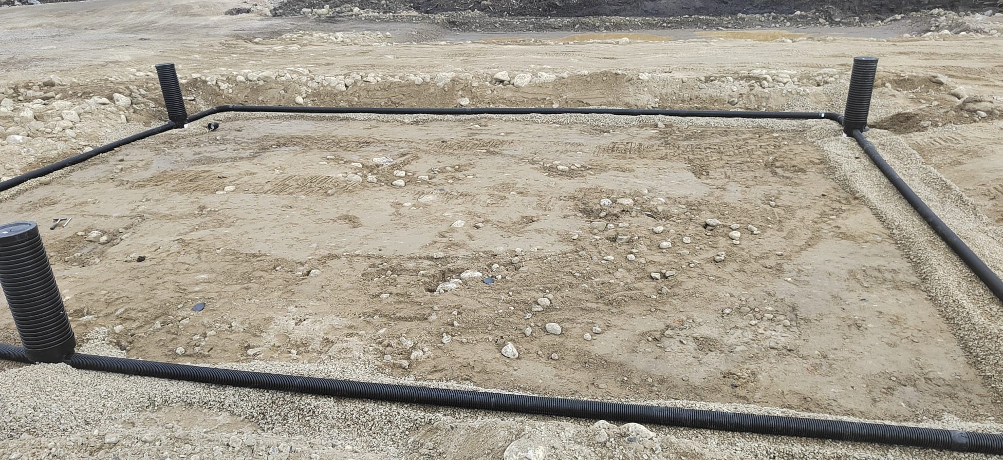 Suorakulmion muotoinen salaojitusjärjestelmä hiekkaisella alueella, jossa on mustat putket ja pystysuorat tarkastuskaivot. Valoisa ja aurinkoinen sää.