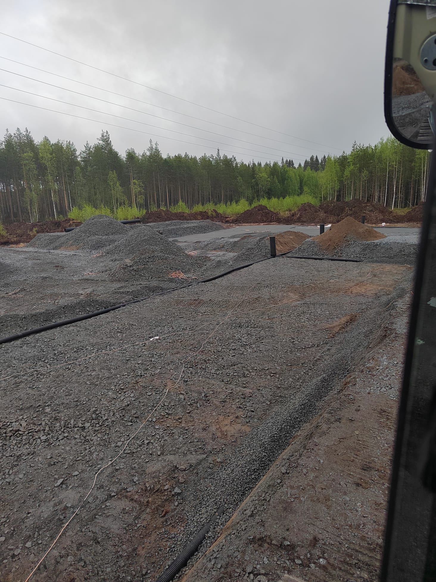 Kuvassa näkyy sorakenttä, jossa on mustia putkia ja sähköjohtoja, taustalla metsä ja harmaa taivas.