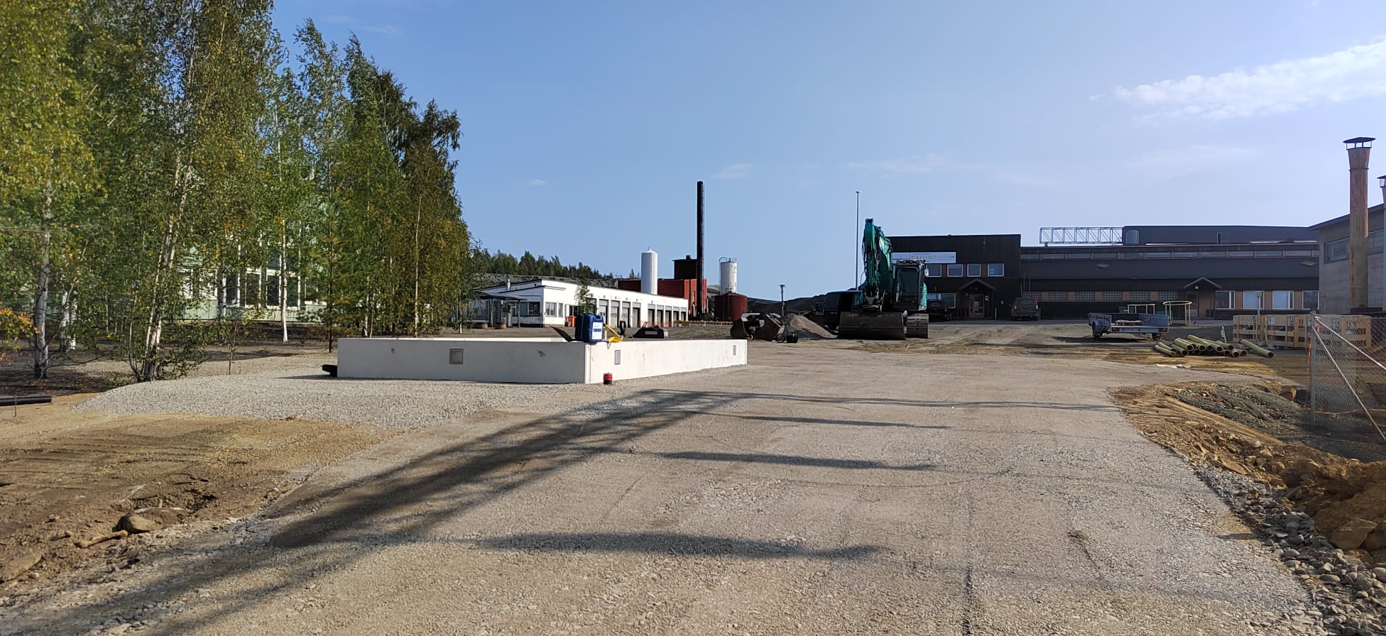 Ulkonäkymä teollisuusalueelle, jossa on rakennuksia, puita, kaivinkone ja avoin asfalttikenttä aurinkoisena päivänä.
