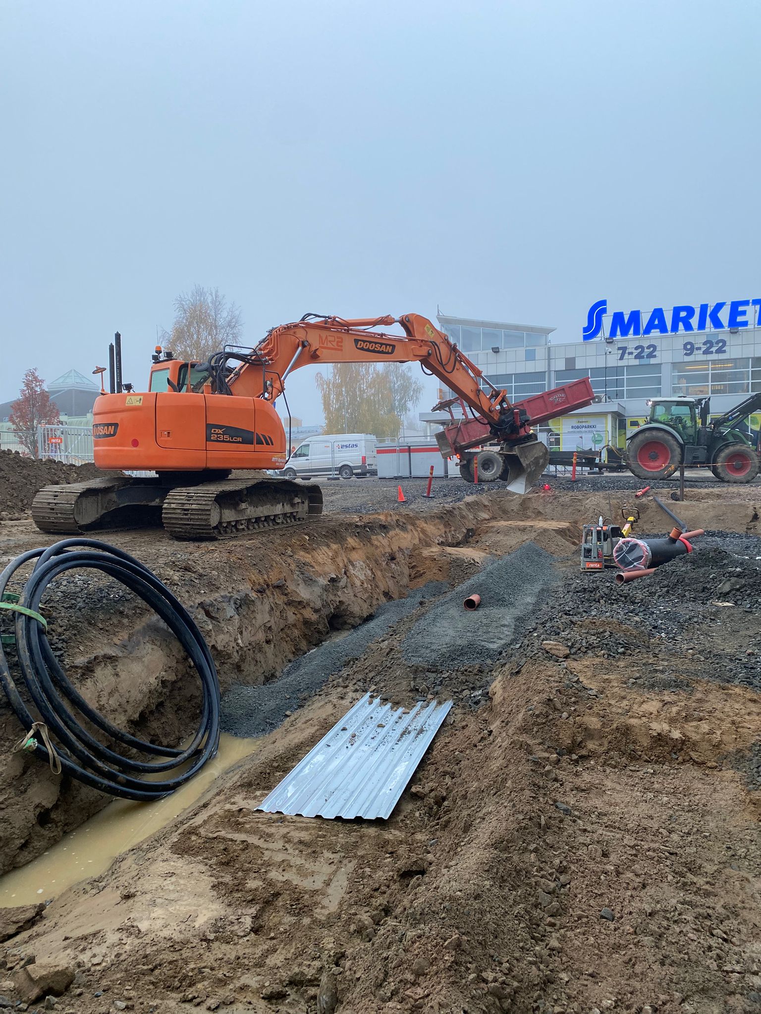 Rakennustyömaa, jossa on oranssi kaivinkone, traktori ja kaivanto, jossa on mustia kaapeleita ja vettä, taustalla S-market rakennus.