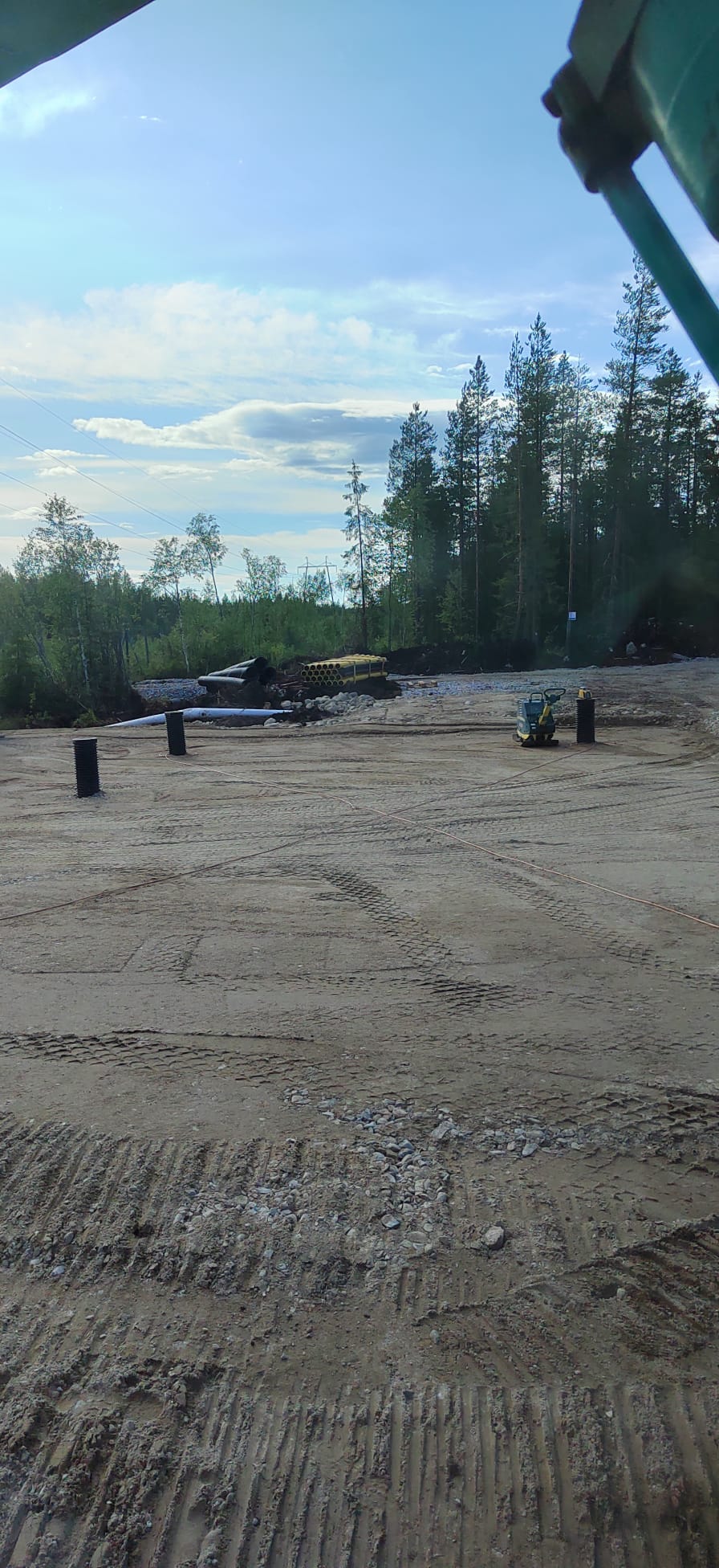 Maanrakennustyömaa, jossa on mustia putkia, keltainen tärytin ja puita taustalla, valoisa sää.