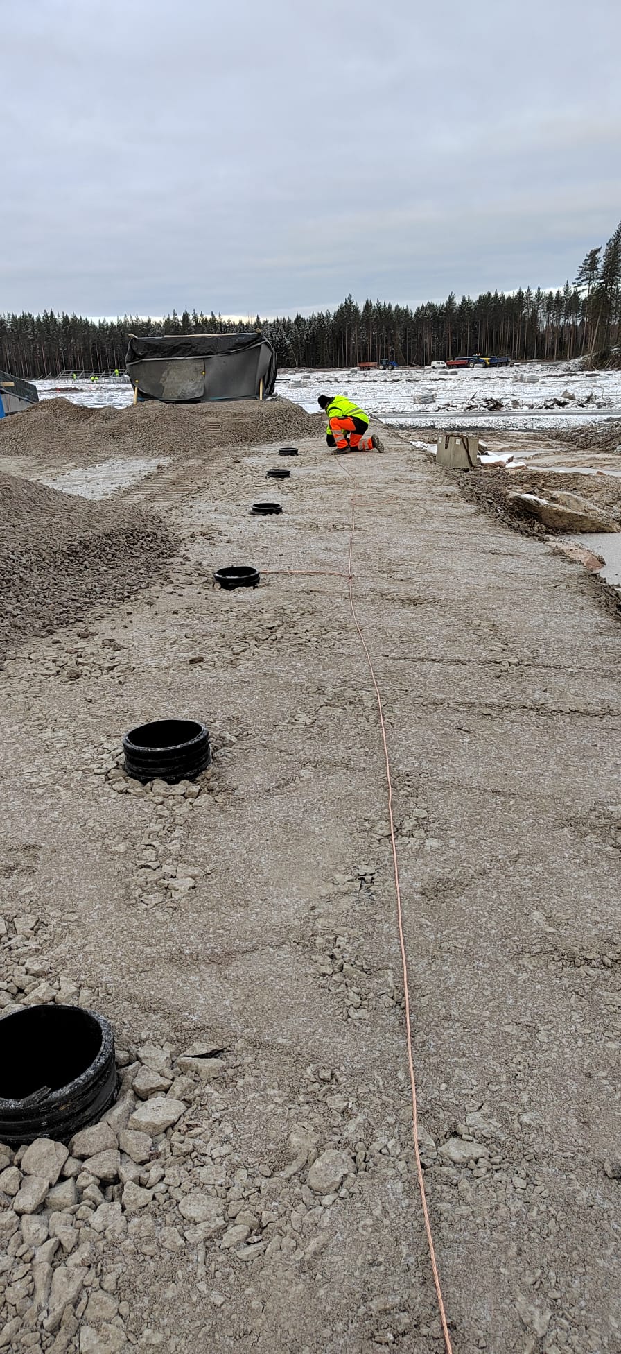 Ulkona työmaalla on mies huomiovaatteissa, joka työskentelee putkien parissa, taustalla näkyy metsää ja taivas on pilvinen.